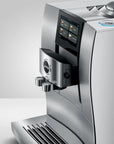 A close-up studio shot of a stainless steel Jura coffee machine, showing the front display screen, water dispenser spout, drip tray, and part of the bean hopper against a clean light gray background.