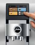 A close-up of a Jura Z10 coffee machine with a human hand touching the touchscreen display, set against a brushed stainless steel background.