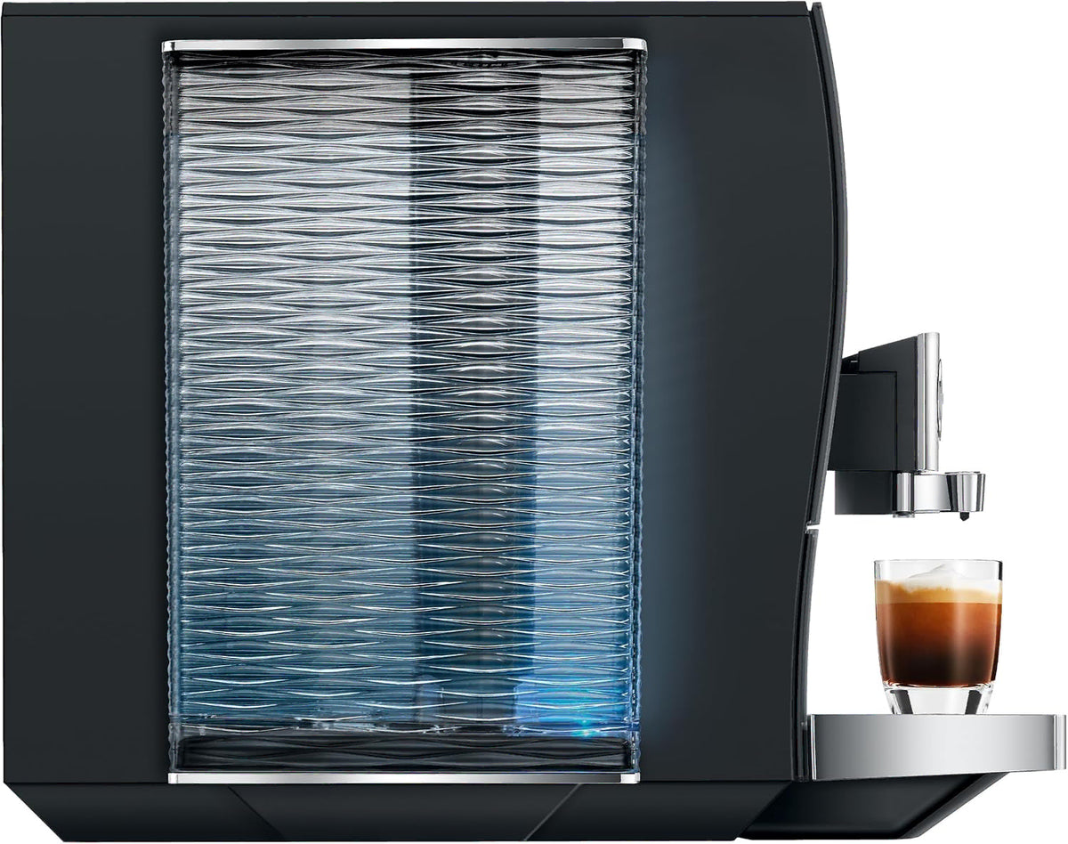 Side view of a sleek black coffee machine with a large ribbed transparent water tank and a small glass of espresso on the drip tray, shown against a plain white background.