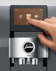A close-up of a Jura Z10 coffee machine with a human hand touching the touchscreen interface, set against a clean gray background.