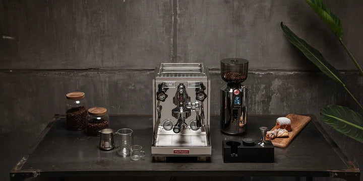 A professional espresso machine with a stainless steel portafilter and a dark coffee grinder, displayed on a dark table against a textured gray wall with coffee beans and a small pastry.