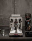 A professional espresso machine with a stainless steel portafilter and a dark coffee grinder, displayed on a dark table against a textured gray wall with coffee beans and a small pastry.
