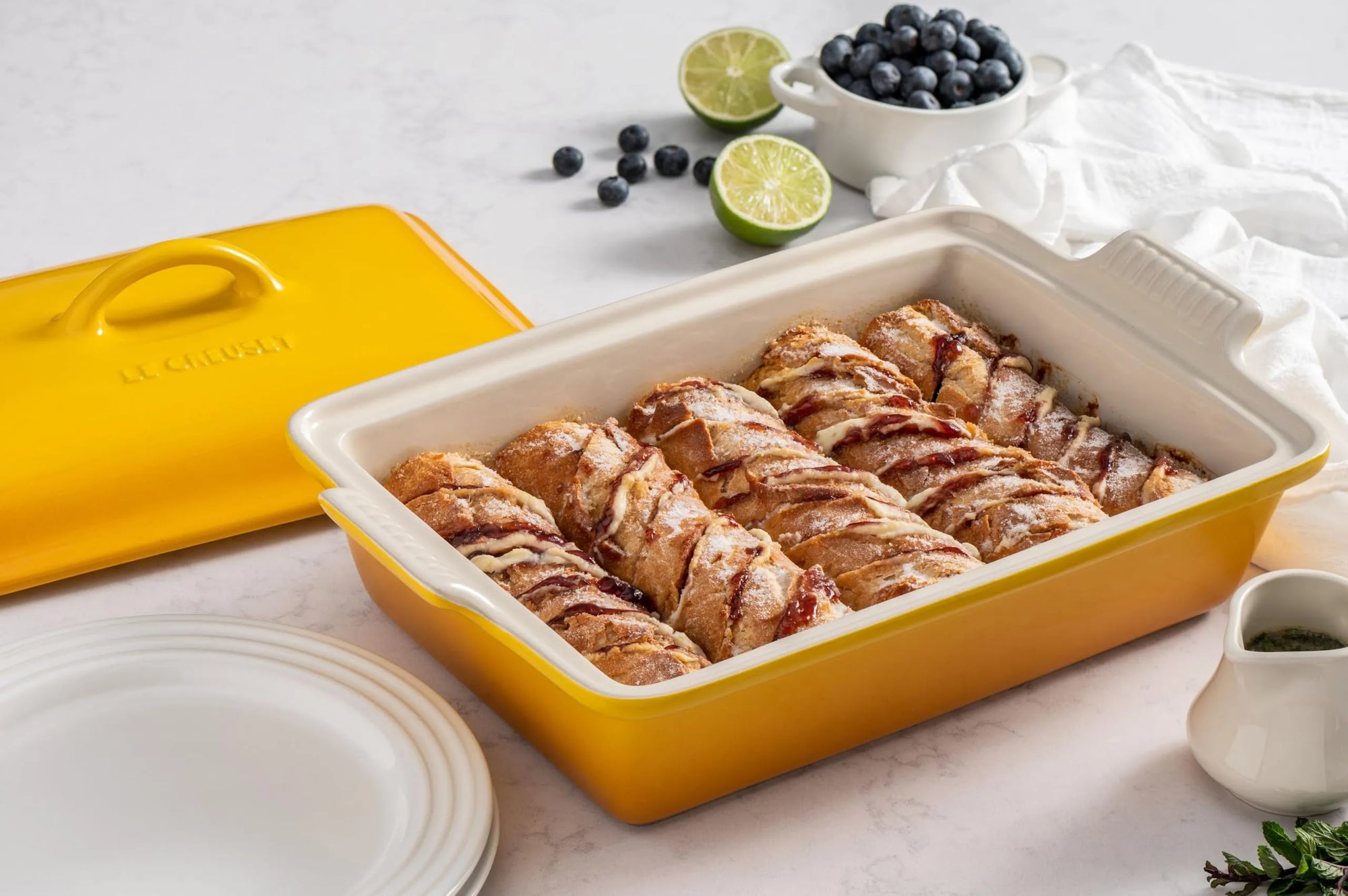 A yellow ceramic baking dish with a cream interior, filled with baked cinnamon rolls, shown with its matching yellow lid on a light marble surface. Blueberries, limes, plates, and a small white pitcher are styled around it.