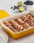 A yellow ceramic baking dish with a cream interior, filled with baked cinnamon rolls, shown with its matching yellow lid on a light marble surface. Blueberries, limes, plates, and a small white pitcher are styled around it.