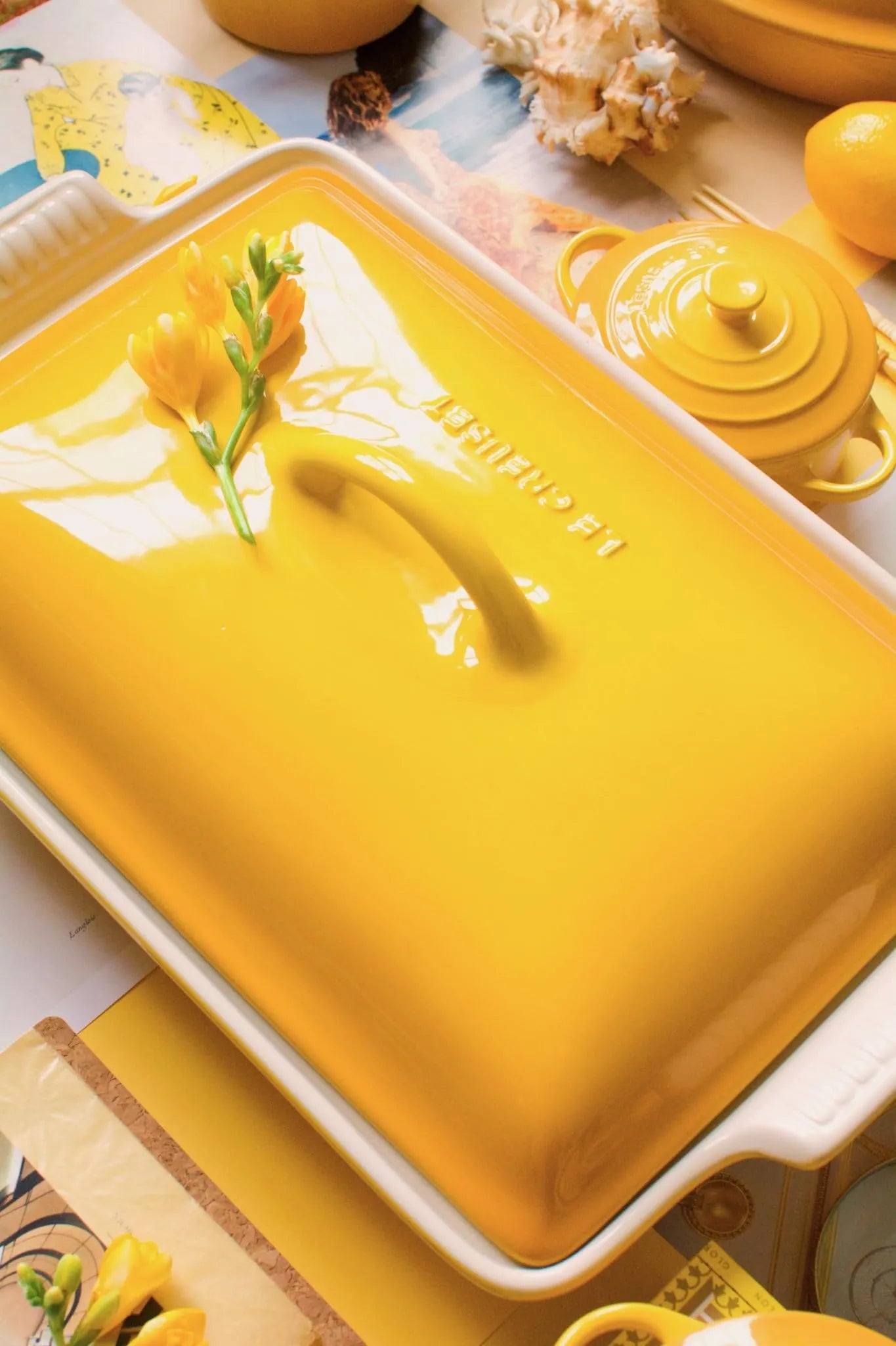 A bright yellow ceramic casserole dish with a matching lid, photographed in a styled tabletop setting with other yellow cookware and small floral accents.