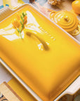 A bright yellow ceramic casserole dish with a matching lid, photographed in a styled tabletop setting with other yellow cookware and small floral accents.