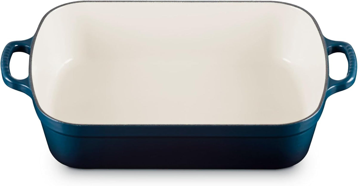 A rectangular enameled baking dish with a deep blue exterior and a cream-colored interior, featuring two side handles.
