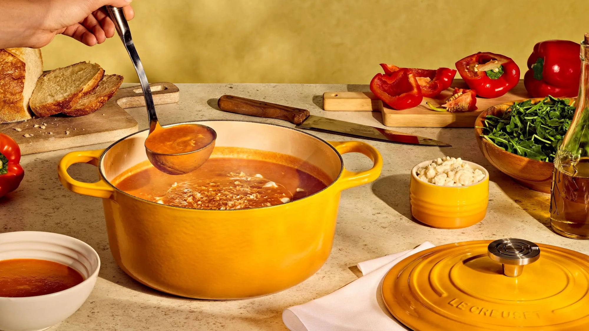 A yellow Le Creuset Dutch oven filled with orange-red soup, with a hand using a ladle to scoop soup. The scene is set on a kitchen counter surrounded by ingredients like bread, red peppers, greens, and a bowl of cheese, with the matching lid resting nearby.