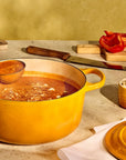 A yellow Le Creuset Dutch oven filled with orange-red soup, with a hand using a ladle to scoop soup. The scene is set on a kitchen counter surrounded by ingredients like bread, red peppers, greens, and a bowl of cheese, with the matching lid resting nearby.