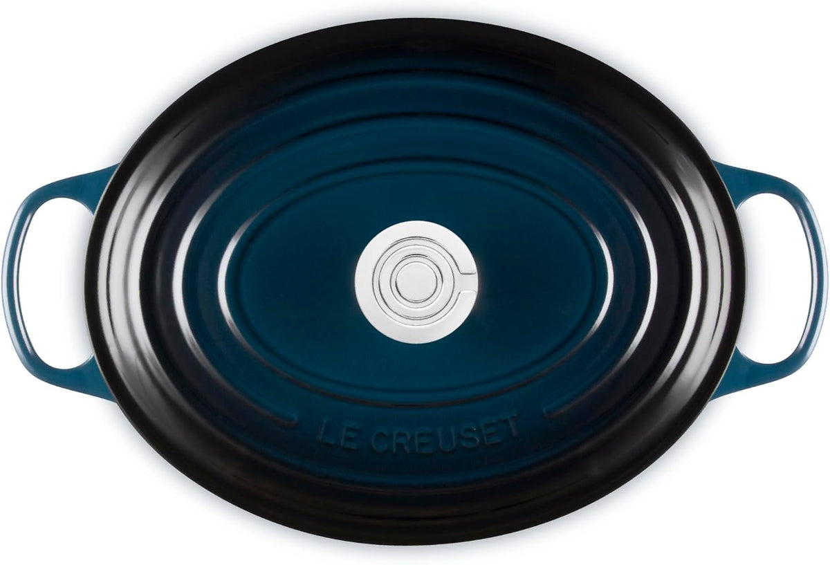 Top view of an oval Le Creuset enameled cast iron Dutch oven with a dark teal blue finish and black rim, featuring side handles and a silver-colored center knob.