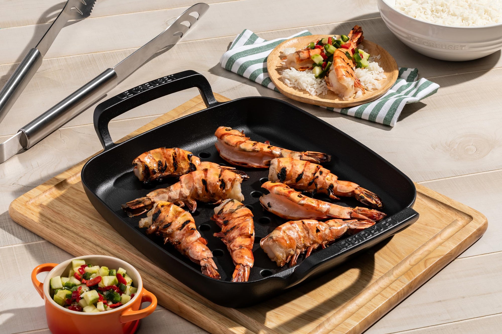 Grilled shrimp on a black grill pan with a wooden cutting board, knife, and bowls of salad and rice.