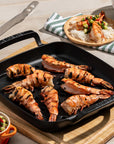 Grilled shrimp on a black grill pan with a wooden cutting board, knife, and bowls of salad and rice.