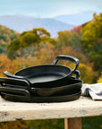Black cast iron pans on a wooden table with a scenic background of mountains and trees.