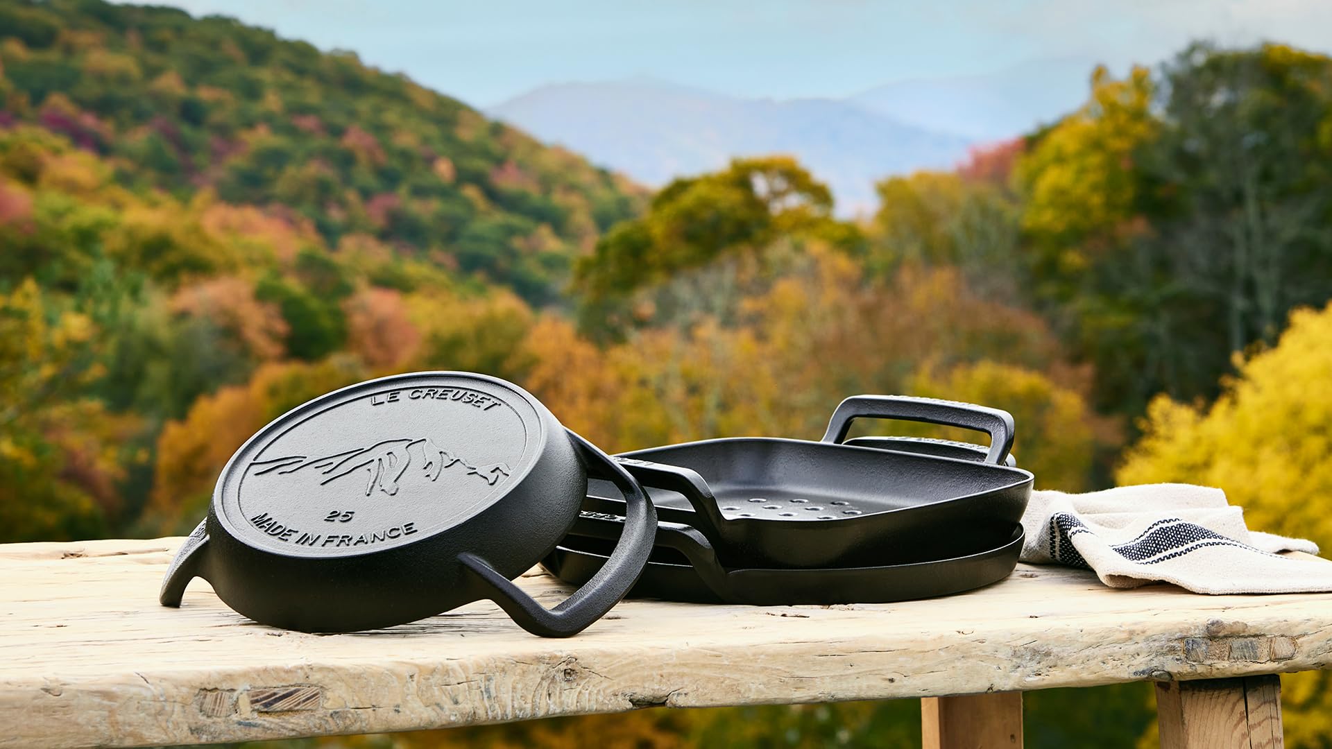 Three cast iron skillets on a wooden table with a scenic background of trees and mountains.