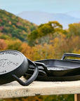 Three cast iron skillets on a wooden table with a scenic background of trees and mountains.