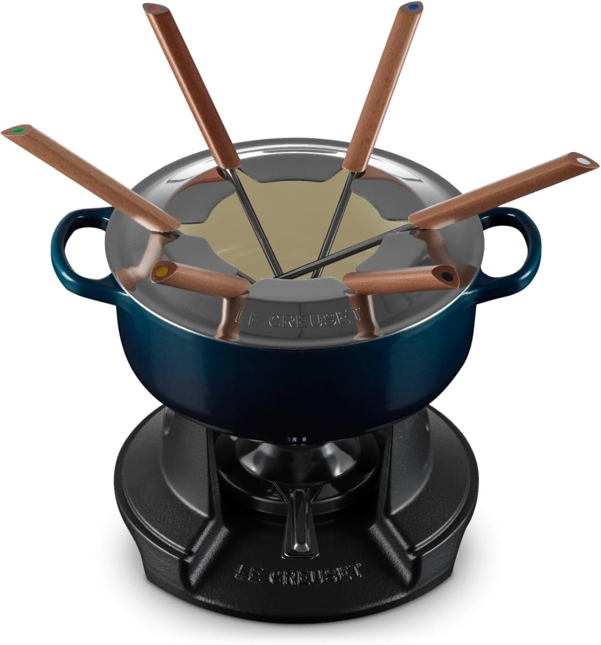 Le Creuset fondue set featuring a dark blue enameled cast iron pot on a black cast iron burner base, with a stainless steel lid, six copper-colored fondue forks, and a beige fondue dip, all shown on a white background.