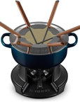 Le Creuset fondue set featuring a dark blue enameled cast iron pot on a black cast iron burner base, with a stainless steel lid, six copper-colored fondue forks, and a beige fondue dip, all shown on a white background.