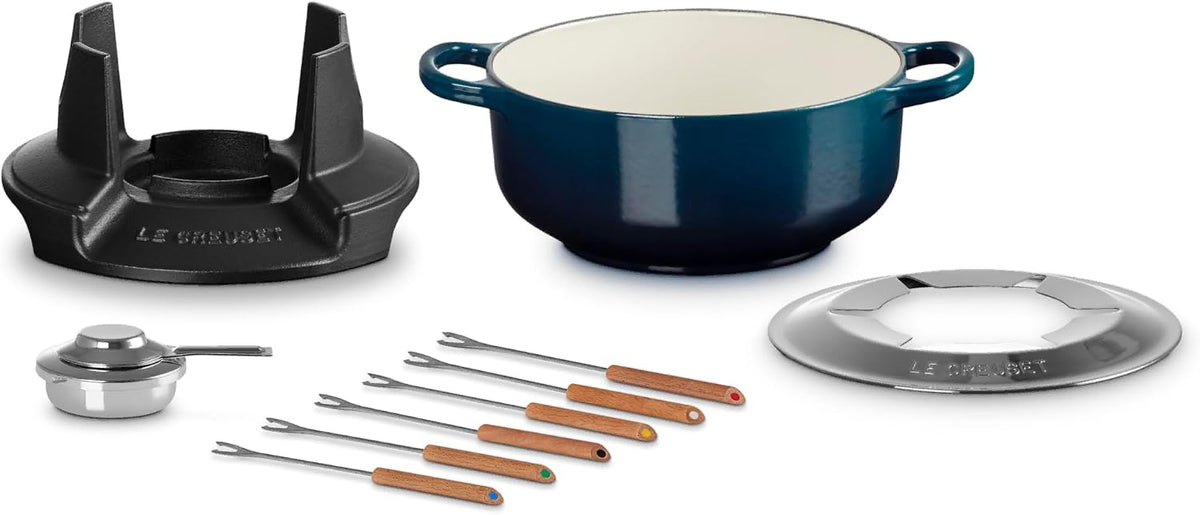 Le Creuset cast iron cookware set including a navy blue enameled pot, black burner stand, stainless steel lid, and six wooden-handled utensils on a white background.