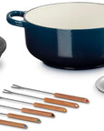 Le Creuset cast iron cookware set including a navy blue enameled pot, black burner stand, stainless steel lid, and six wooden-handled utensils on a white background.