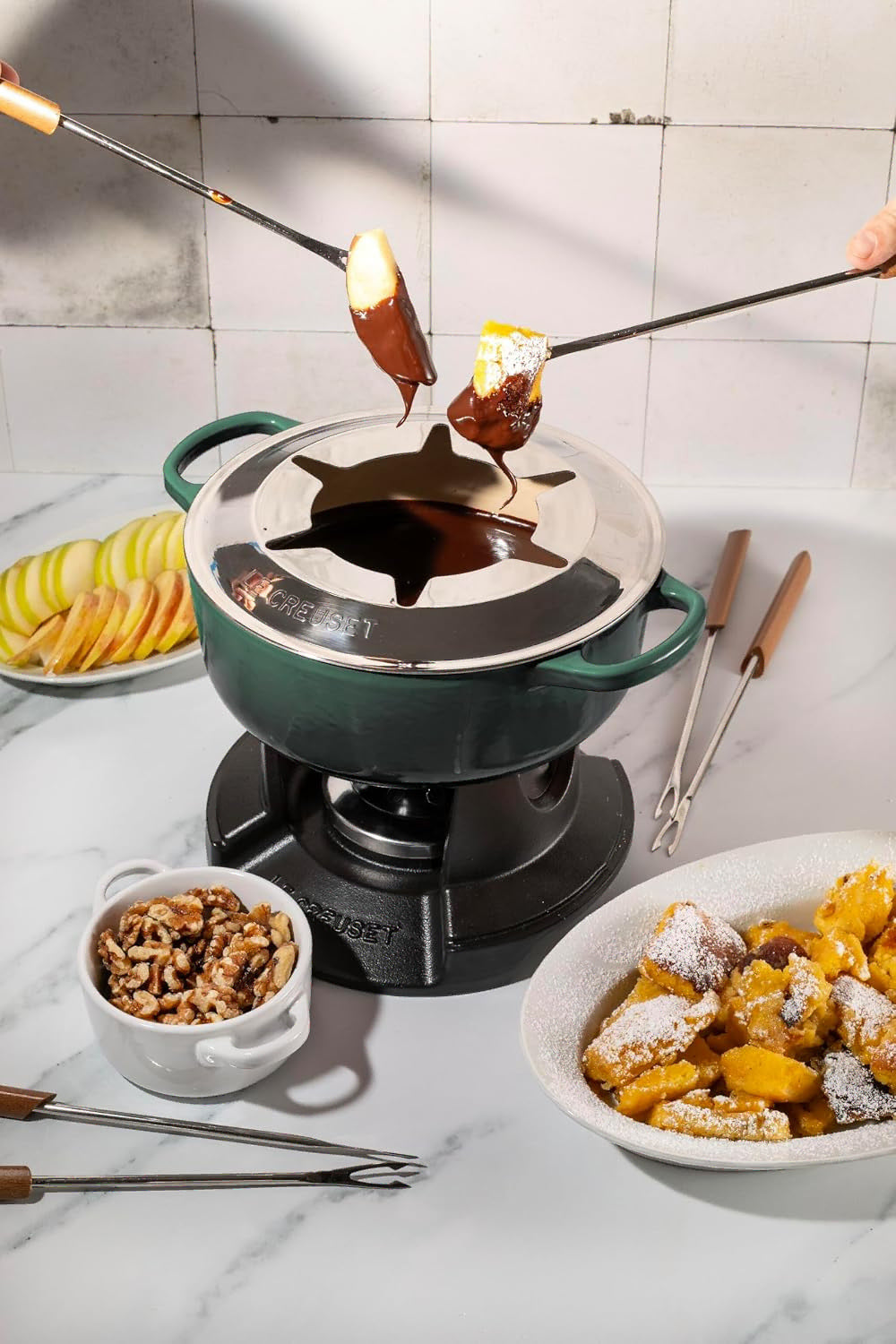 A star-shaped fondue pot on a portable burner with chocolate-dipped food being lifted by two hands using long-handled forks. The pot is dark green and black, set on a white marble countertop with plates of fruit, nuts, and dessert in a kitchen setting.