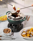 A star-shaped fondue pot on a portable burner with chocolate-dipped food being lifted by two hands using long-handled forks. The pot is dark green and black, set on a white marble countertop with plates of fruit, nuts, and dessert in a kitchen setting.