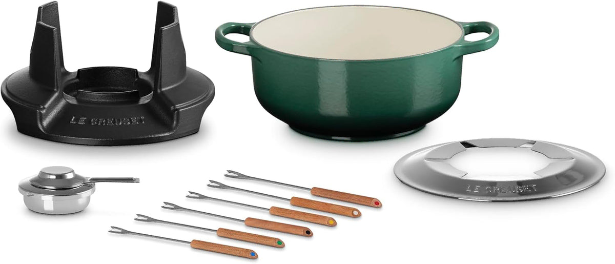 Le Creuset Dutch oven set on a white background, featuring a dark green enameled cast iron pot with a cream interior, a black burner stand, a silver lid, a small silver pot insert, and six metal skillets with wooden handles.
