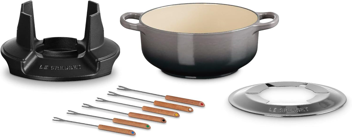 Le Creuset cast iron pot set with a dark gray to black ombre finish, cream interior, matching lid, and six copper utensils with wooden handles on a white background.