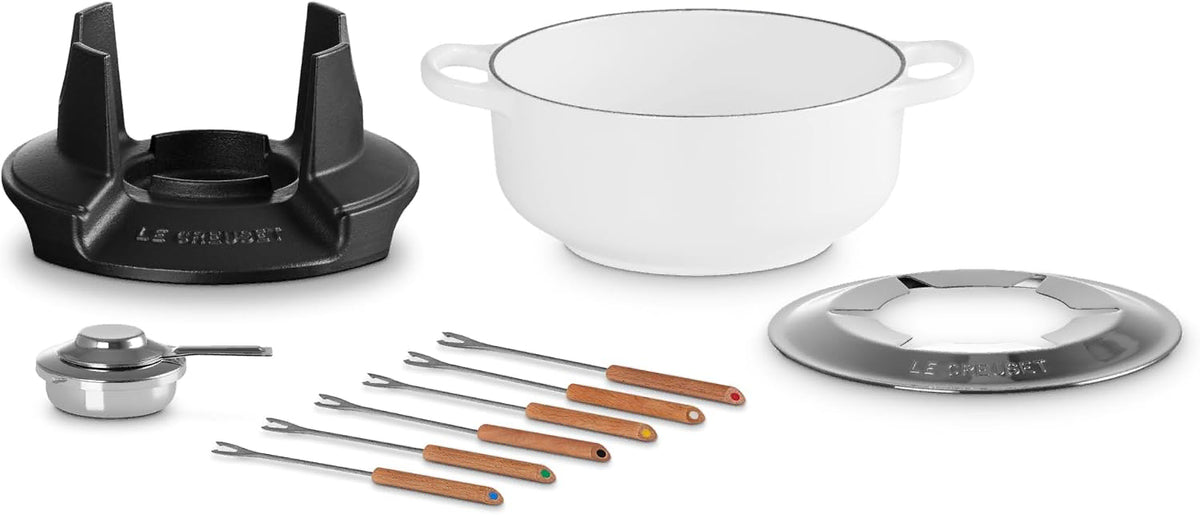 Le Creuset cast iron cookware set on a white background, featuring a white enameled pot with a cream interior, a black cast iron burner grate, a small silver saucepan, and multiple stainless steel skewers with wooden handles.
