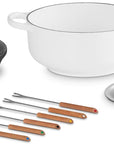 Le Creuset cast iron cookware set on a white background, featuring a white enameled pot with a cream interior, a black cast iron burner grate, a small silver saucepan, and multiple stainless steel skewers with wooden handles.