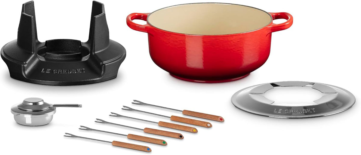 Le Creuset cast iron cookware set on a white background, featuring a red enameled pot with a cream interior, a black cast iron burner grate, a small silver saucepan, and multiple stainless steel skewers with wooden handles.