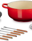 Le Creuset cast iron cookware set on a white background, featuring a red enameled pot with a cream interior, a black cast iron burner grate, a small silver saucepan, and multiple stainless steel skewers with wooden handles.