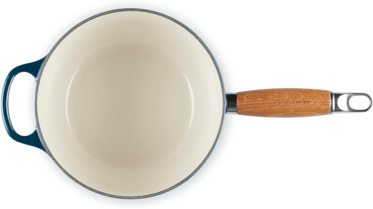 An overhead view of a Le Creuset saucepan with a cream-colored enamel interior, a dark teal blue exterior handle, and a light brown wooden handle with a silver metal loop.