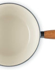 An overhead view of a Le Creuset saucepan with a cream-colored enamel interior, a dark teal blue exterior handle, and a light brown wooden handle with a silver metal loop.