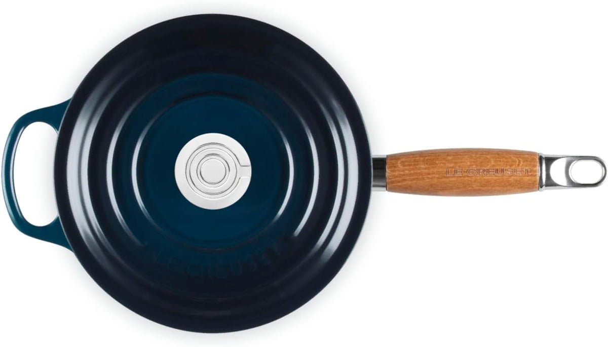 A top-down view of a Le Creuset cast iron skillet with a dark navy blue interior, a teal exterior edge, a light brown wooden handle, and a silver metal loop.