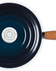 A top-down view of a Le Creuset cast iron skillet with a dark navy blue interior, a teal exterior edge, a light brown wooden handle, and a silver metal loop.