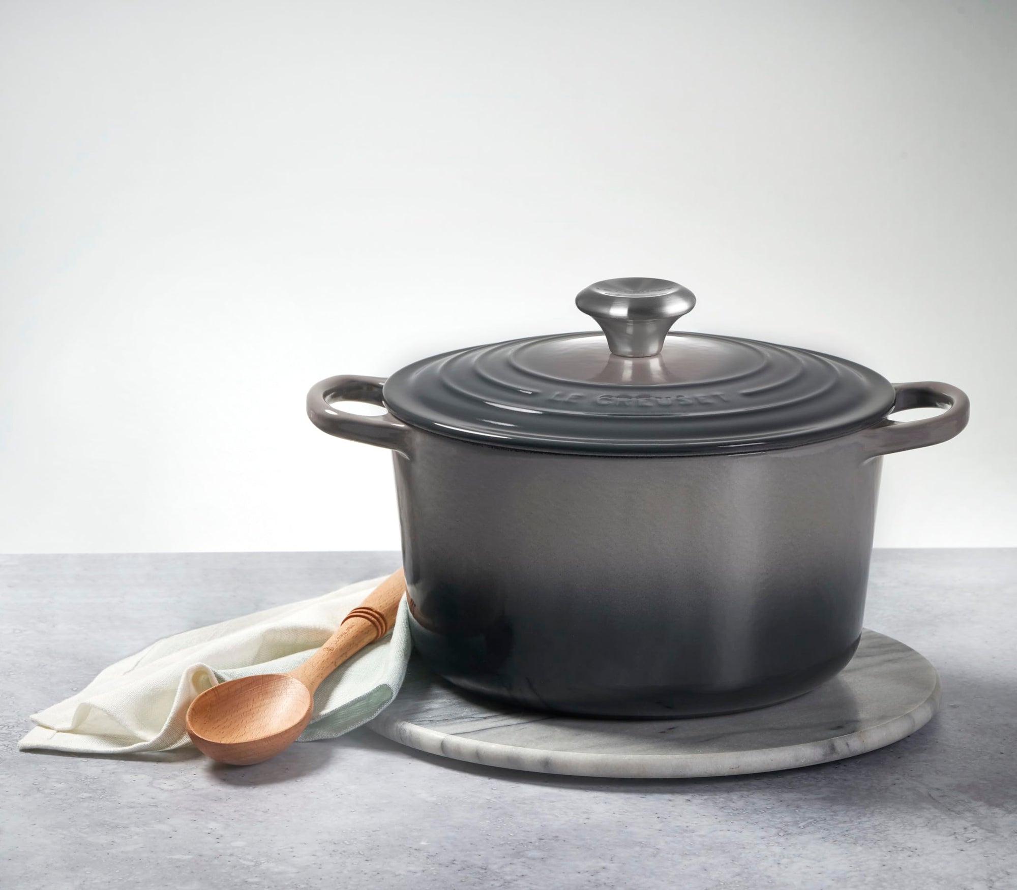 Oyster cast iron round oven with handles and lid on a marble trivet on a countertop with wooden spoon and napkin