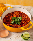 A yellow Le Creuset enameled cast iron pot filled with a red chili stew, surrounded by bowls of onions, lime, avocado, and tortillas on a beige stone countertop.