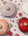 White, Peche, Cerise, and chiffon pink heart shaped 1.25 qt cocotte with handles and a light gold heart knob next to heart shaped candy and flower petals on a marble countertop
