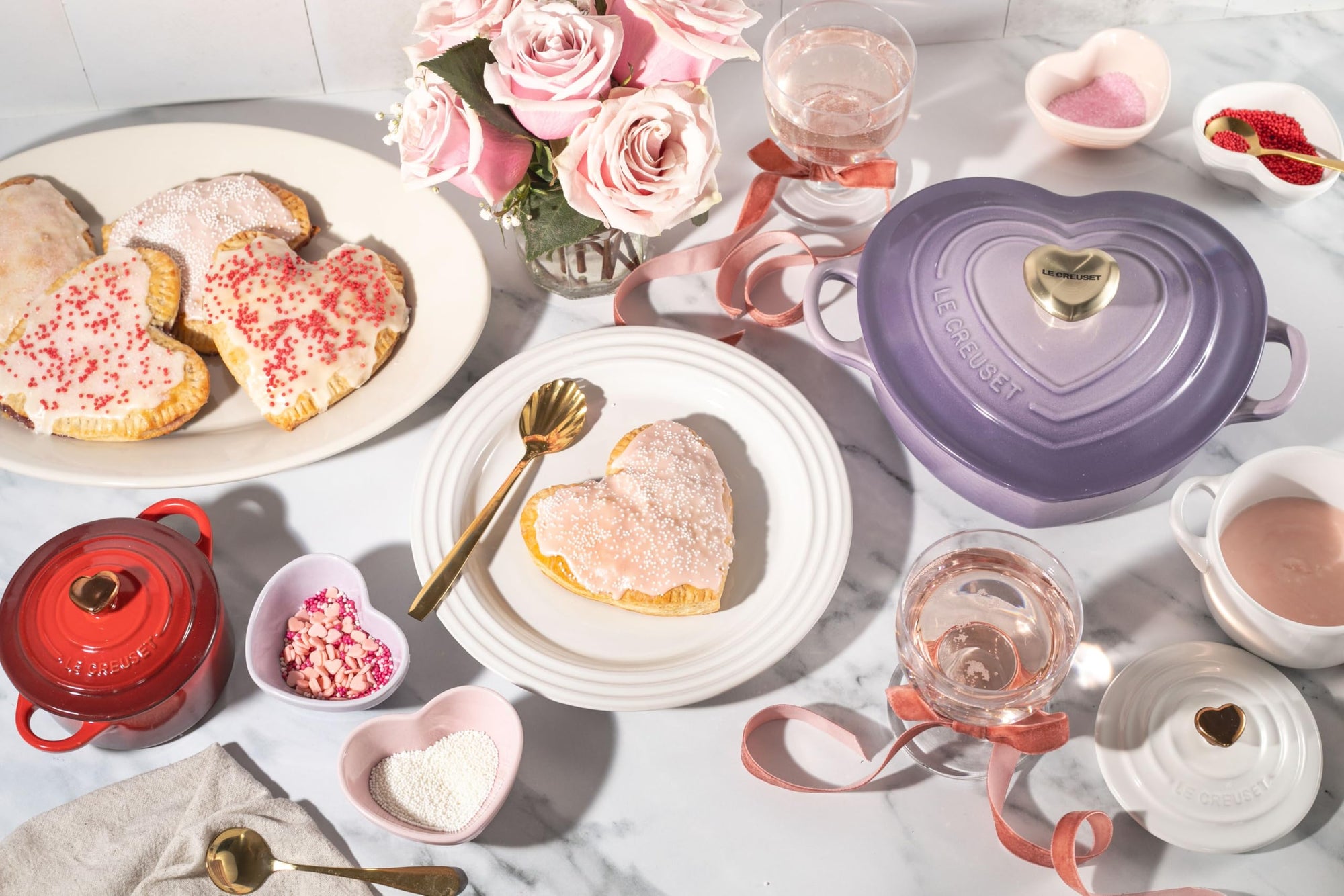 Provence heart shaped 1.25 qt cocotte with handles and a light gold heart knob next to heart shaped sugar cookies, baking supplies for cookies, and pink roses on a marble countertop