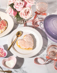 Provence heart shaped 1.25 qt cocotte with handles and a light gold heart knob next to heart shaped sugar cookies, baking supplies for cookies, and pink roses on a marble countertop