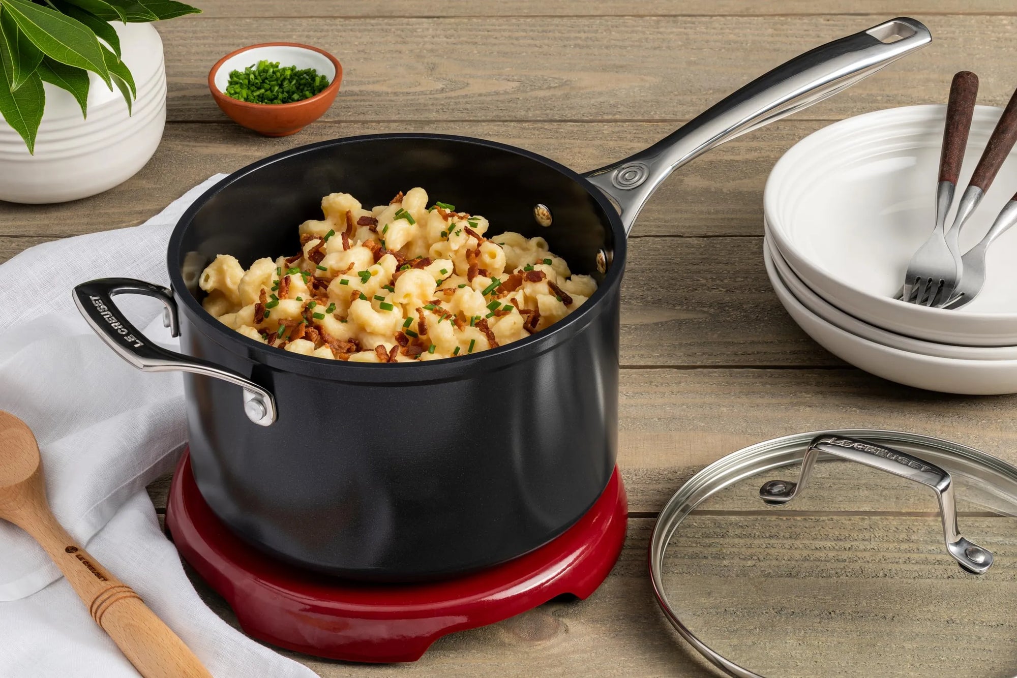 Le Creuset Essential Non-stick Ceramic 3 qt Saucepan with Lid with cooked mac and cheese next to bowls and forks on a wooden surface
