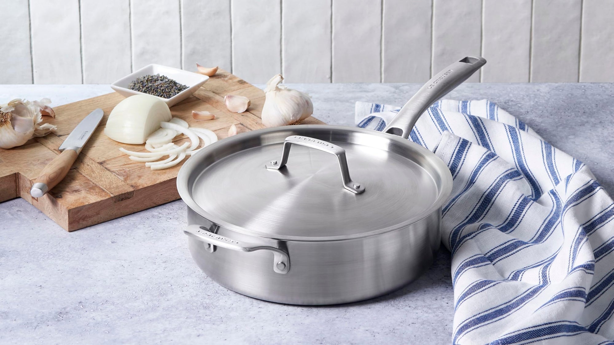 Le Creuset stainless steel pot with handles and lid next to blue and white tablecloth and a cutting board with garlic, onion, and knife on a blue marble countertop