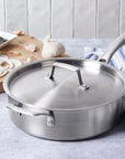 Le Creuset stainless steel pot with handles and lid next to blue and white tablecloth and a cutting board with garlic, onion, and knife on a blue marble countertop