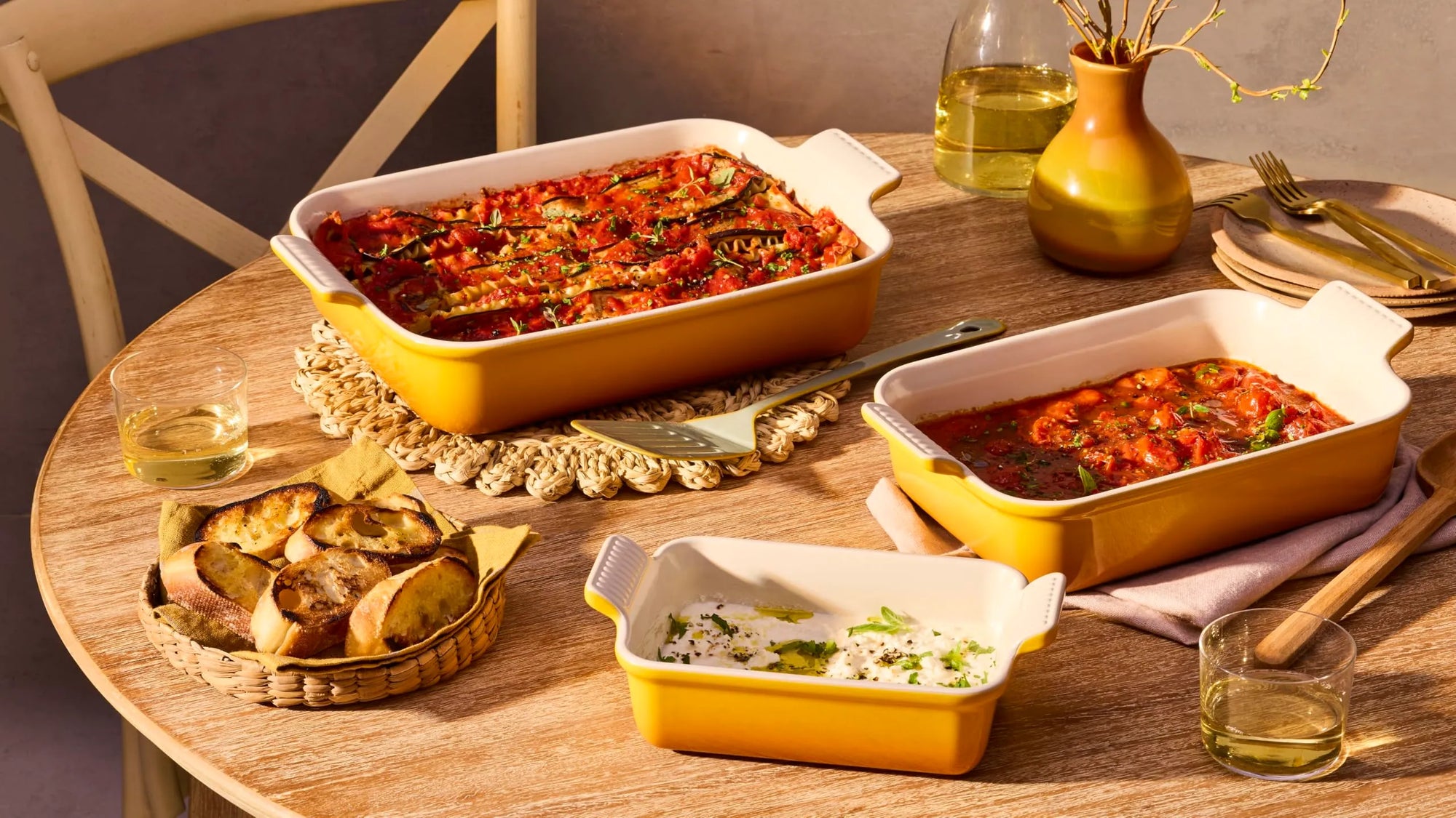 Three yellow and white ceramic baking dishes filled with cooked pasta and side dishes, arranged on a wooden dining table with bread, glasses, and tableware in a warm indoor setting.