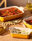 Three yellow and white ceramic baking dishes filled with cooked pasta and side dishes, arranged on a wooden dining table with bread, glasses, and tableware in a warm indoor setting.