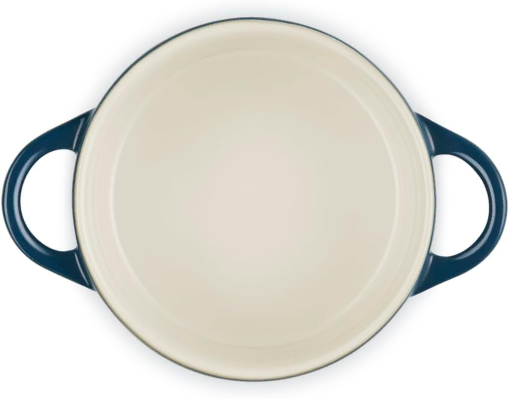 An oval-shaped enameled cast iron Dutch oven shown from a top-down perspective, featuring a cream-colored interior and dark navy blue handles.