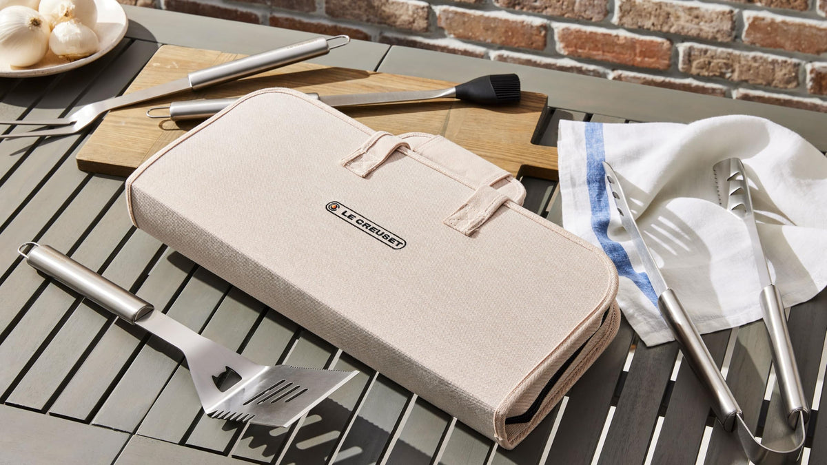 Beige carrying case for grill tools on a table with utensils and a brick wall background