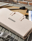 Beige carrying case for grill tools on a table with utensils and a brick wall background