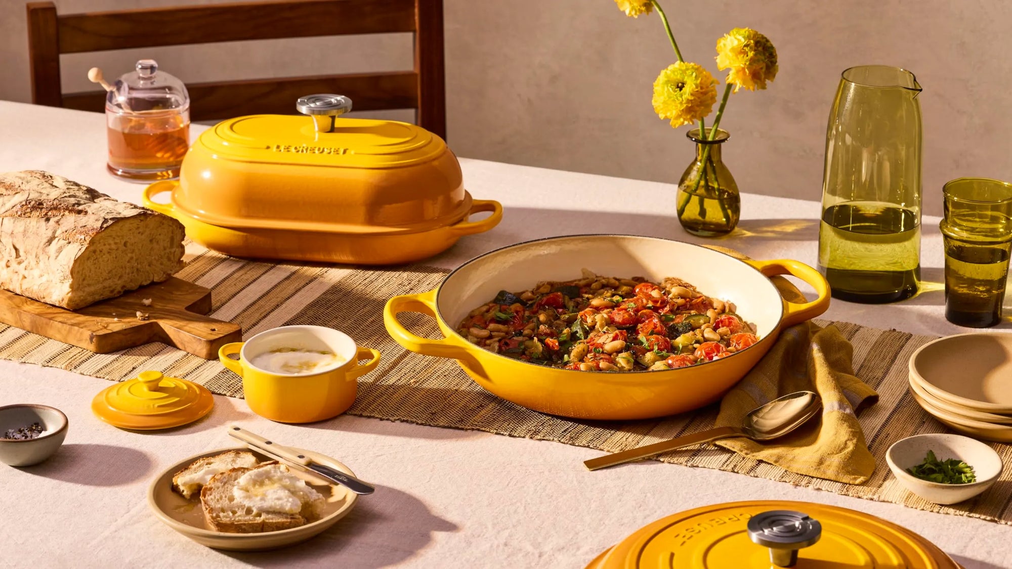 A yellow enameled cast iron casserole dish with a matching lid sits on a dining table, surrounded by a breakfast spread including bread, a bowl of soup, a glass pitcher, and yellow flowers in the background.