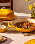 A yellow enameled cast iron casserole dish with a matching lid sits on a dining table, surrounded by a breakfast spread including bread, a bowl of soup, a glass pitcher, and yellow flowers in the background.